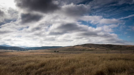 A golden field stretches to meet rolling hills under a dramatic, cloudy sky.