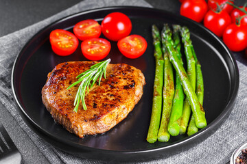 Close up, macro of tasty grilled beef steak served with green asparagus and cherry tomatoes on black plate, with cutlery. Perfect for food menu, diet, or culinary design concepts.