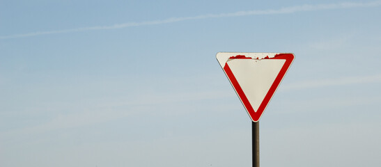 Old and partially damaged yield road sign (give way) with peeling paint, isolated on a pole against a bright blue sky. Symbol of traffic regulation, caution, road safety, and minimalism