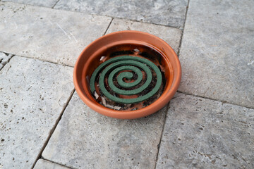 A green mosquito coil resting in a terracotta dish on a natural stone surface. The spiral shape of...