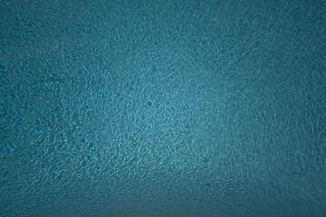 Abstract image of a gently rippled blue water surface, likely from a swimming pool. The light...