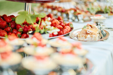 Close-up of fresh strawberries arranged on tiered silver trays with delicate desserts on a white tablecloth in outdoor sunlight, evoking a vibrant summer celebration