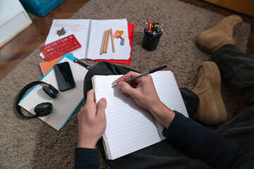 Student writing in an open notebook sitting on a rug on the floor of his room.