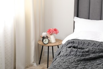Bedside table with alarm clock, flowers and books near bed indoors
