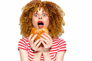 Surprised young woman with curly hair holding a hamburger, wearing a striped shirt, expressing...
