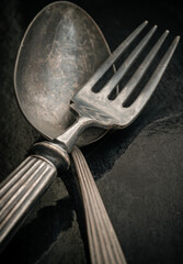 Ancient worn vintage cutlery on the black background. Crossed black metal spoon and fork in dark...