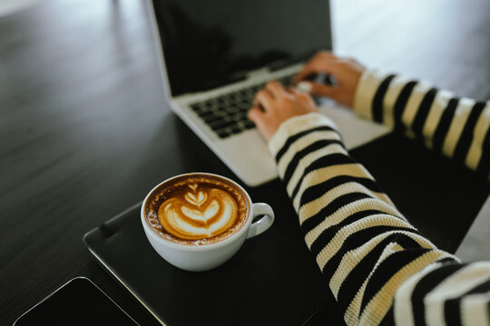 Young Asian woman making latte in a relaxed atmosphere with laptop and notebook and notepad while combining work and relaxation during a quiet coffee break at home or office.