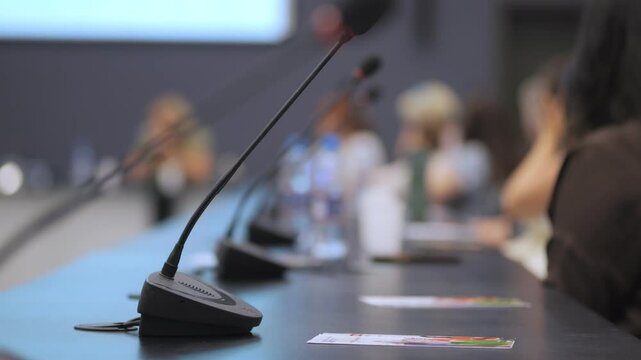 Microphone sits on a table with a few other microphones. The table is surrounded by people, some of whom are talking on cell phones. The scene suggests a professional setting
