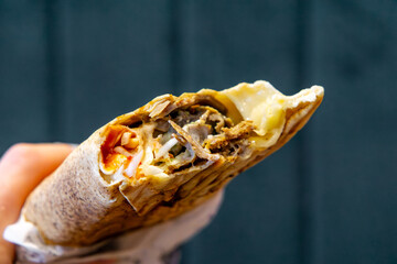 hand holding a wrap kebab with visible meat, vegetables, and melted cheese. Dark blurred background...