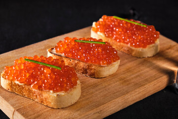 Three sandwiches with red caviar on a saucer on wooden plate. Gourmet appetizer of trout caviar on a french baguette slice with butter. Salted salmon roe for fish delicacy concept. Front view.
