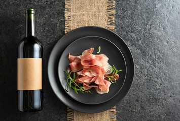 Bottle of red wine and prosciutto on a black plate.