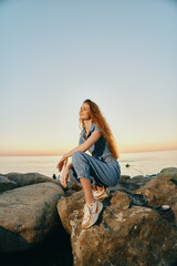 Lifestyle woman in modern boho-western denim street style sitting on rocks near the sea during...