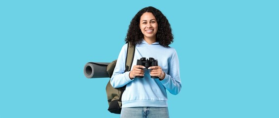 Young African-American female tourist with backpack and binoculars on blue background