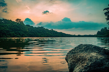 Serene lake view at sunset, framed by large rock and lush green trees with colorful reflections on still water. Perfect for tranquil nature scenes, eco tourism themes, and peaceful landscapes