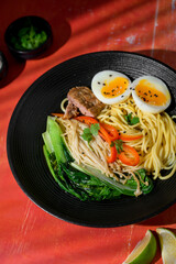 Top view of ramen in black bowl red background. Traditional Japanese noodle soup with rich broth, vegetables, and toppings.