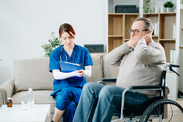 A caring nurse in blue scrubs conducts a neck pain consultation with a senior wheelchair patient at...