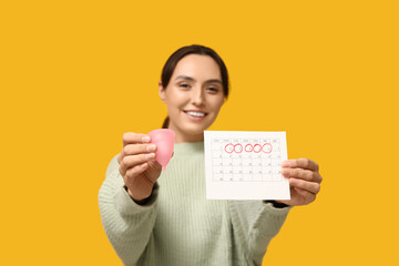 Young woman holding menstrual cup and calendar with marked days of menstruation on yellow background
