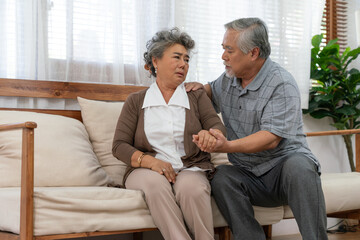 Asian senior couple feeling sad sitting on couch in living room at home. Elderly man consoling and holding his wife hand.