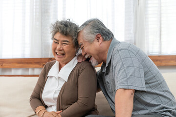 Asian elderly couple enjoying joyful moment together at home, smiling and laughing while sitting closely on a cozy sofa