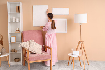 Young woman hanging stylish painting on beige wall in living room, back view
