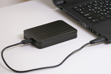 Close-up of an external hard drive connected to a laptop on a white table. Portable storage solution for digital files, data backup, and safe information transfer in modern technology.