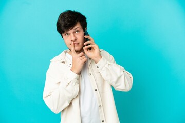 Young Russian man using mobile phone isolated on blue background having doubts while looking up