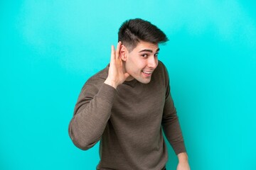 Young caucasian man isolated on blue background listening to something by putting hand on the ear
