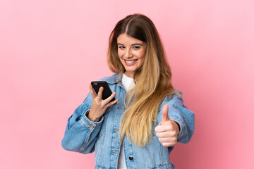 Young blonde woman isolated on blue background using mobile phone while doing thumbs up