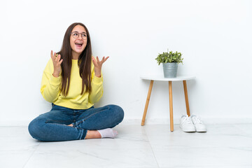 Young Ukrainian woman sitting on the floor of her house isolated on white wall with surprise facial...