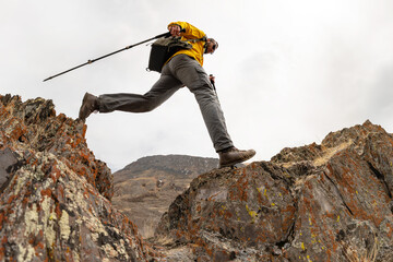 Bottom low angle view of male trekker with backpack and poles jumping over mountain crevice against...