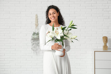 Beautiful young happy African-American woman holding vase with bouquet of white lily flowers near...