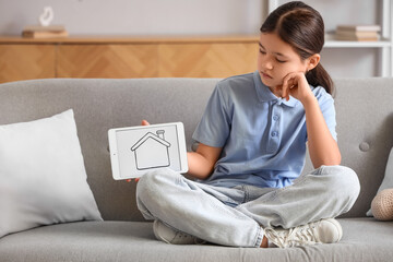 Sad girl holding tablet computer with house figure on sofa at home. Adoption concept