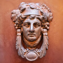 Close-up of an antique bronze door knocker shaped like a woman’s head on a terracotta-colored...