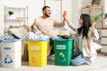 Young couple with sorted rubbish in garbage bins giving high-five to each other at home. Global...