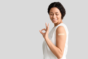Young African-American woman with medical patch showing OK after vaccination on light background