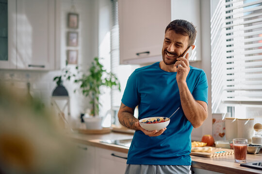 Happy man eating fruit oatmeal while talking on mobile phone in kitchen - Powered by Adobe