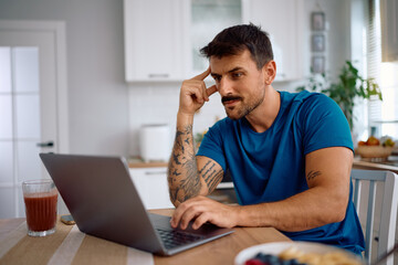 Pensive man reading email on laptop in kitchen.
