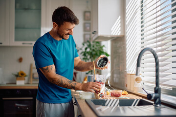 Happy man using blender while preparing fresh fruit smoothie in kitchen.