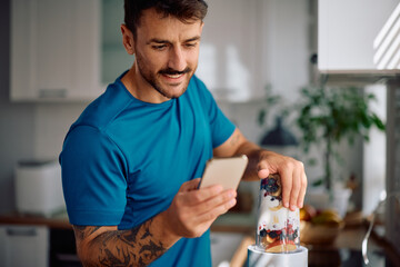 Happy man using cell phone while preparing smoothie in kitchen.