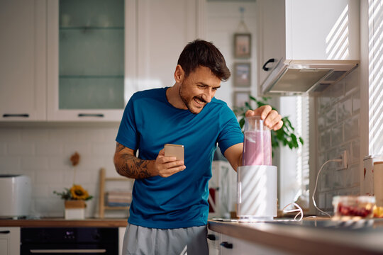 Happy man using mobile phone while preparing smoothie for breakfast at home.