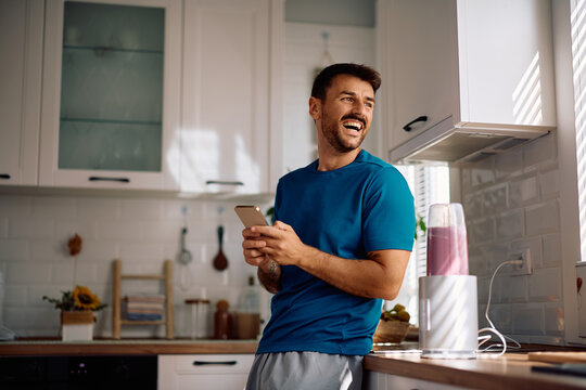 Happy man using cell phone while blending fresh fruit for healthy breakfast in kitchen.