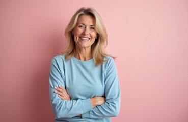 Fototapeta premium Mature woman with blonde hair smiles confidently with arms crossed against pink background. Wears light blue casual t-shirt, conveying friendly, approachable demeanor. Positive expression suggests