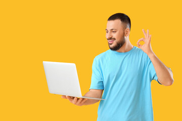 Handsome young man with laptop showing OK gesture on yellow background