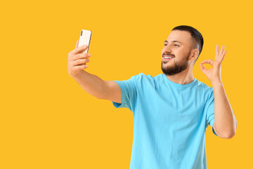 Handsome young man with mobile phone taking selfie while showing OK gesture on yellow background