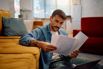 Freelance worker analyzing paperwork while working on laptop at home.