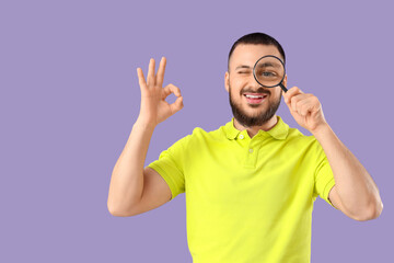 Handsome young man with magnifying glass showing OK gesture on lilac background