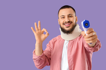 Handsome young man with neck splint and infrared thermometer showing OK gesture on lilac background