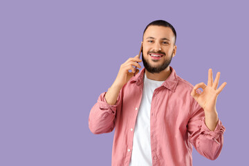 Handsome young man talking by mobile phone and showing OK gesture on lilac background