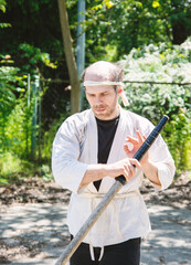 Man in martial arts gi with headband, holding a wooden sword outdoors, focused on preparation....