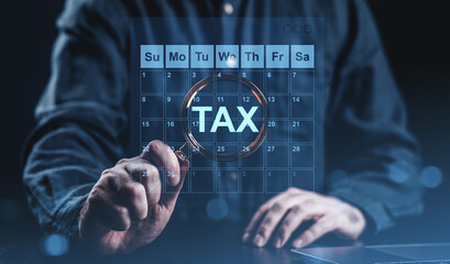 Business person holding magnifying glass over digital calendar with the word TAX highlighted,...